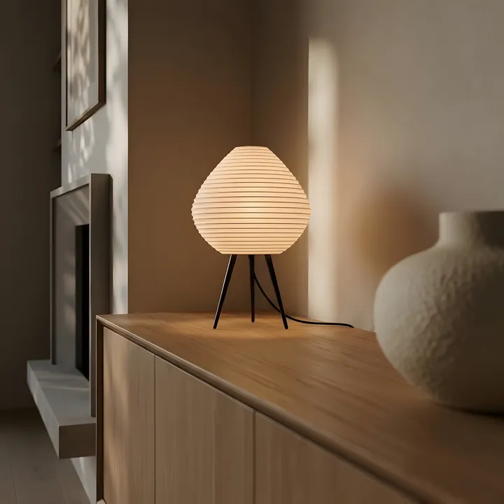 A ridged tripod lamp lit on a wooden sideboard, beside a ceramic vase